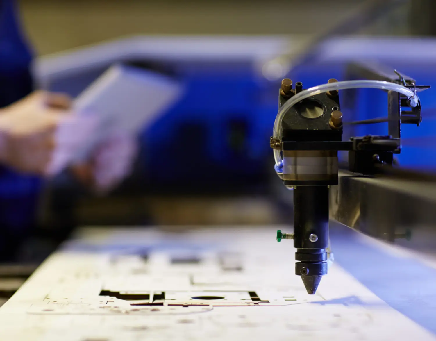 A close-up of a precision cutting tool, poised above a detailed design on a sheet, with a person holding a tablet in the background
